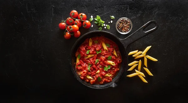 Domates sosu ve fesleğen ile pişirme makarna penne demir döküm tavada servis edilir. Siyah kayrak arka plan, üst görünüm