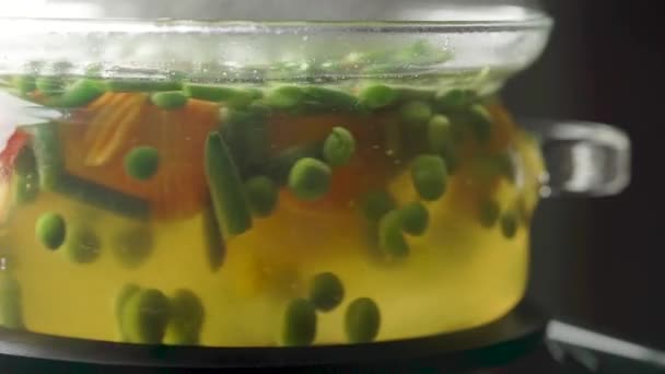 Glass pot with boiling vegetable soup on stove, with foam and bubbles. Side view. — Stock Video