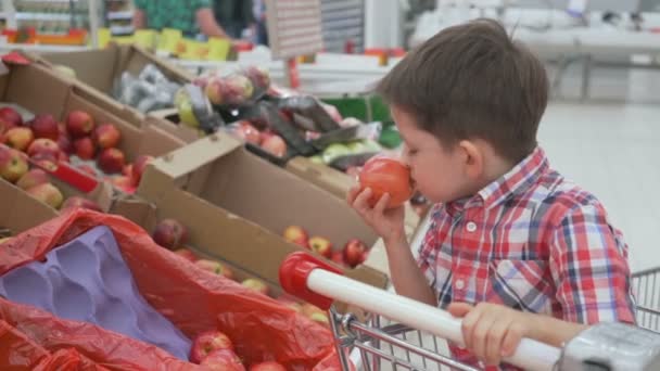 Rolig liten pojke plocka äpple från lådan sitter i vagnen, under familjens shopping i stormarknad — Stockvideo