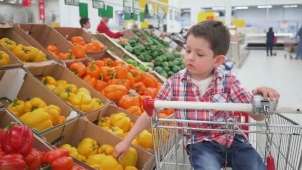 Ragazzino al supermercato che puzza di pepe bulgaro giallo seduto nel carrello. Shopping in negozio, prodotti freschi per la cucina e la cucina . — Video Stock