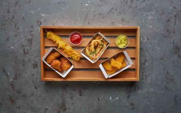 Nuggets de pollo frito, patas, pulpo crujiente y papas fritas en espiral en bandeja de madera, vista superior, espacio para copiar. concepto de comida callejera sobre fondo de piedra —  Fotos de Stock