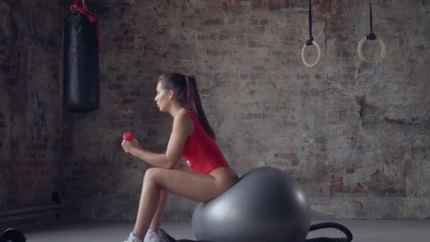 Chica en traje de baño rojo entrenamiento con pesas mientras está sentado en swiss-ball en el fondo de una pared de ladrillo — Vídeos de Stock