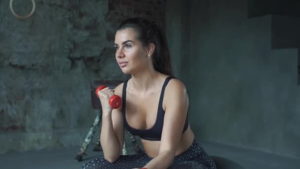 Молодая девушка в красной спортивной одежде делает упражнения с гантелями на фоне кирпичной стены — стоковое видео