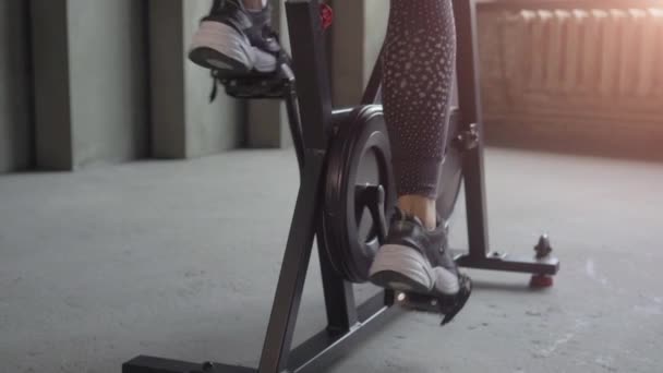 Сексуальная молодая женщина в черной спортивной одежде катается на велосипеде в спортзале и слушает музыку из наушников — стоковое видео