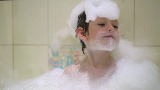 Смешной ребенок играет с водой и пеной в ванной комнате — стоковое видео