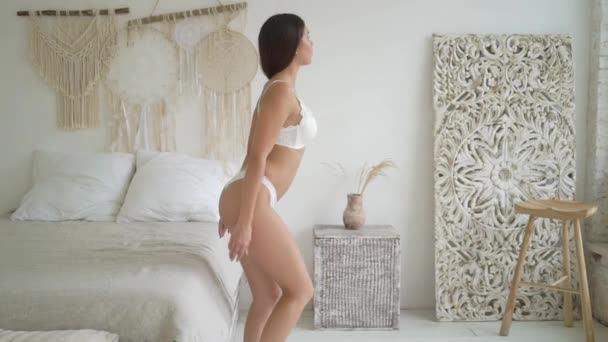 Σέξι μελαχρινή κυρία που ποζάρει σε λευκά εσώρουχα, κάθεται στο κρεβάτι — Αρχείο Βίντεο