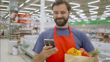 Gülümseyen süpermarket çalışanı cep telefonu kullanıyor ve süpermarkette alışveriş çantası tutuyor.