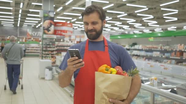 Smiling supermercado empleado utilizando el teléfono móvil y la celebración de la bolsa de compras en el supermercado — Vídeos de Stock