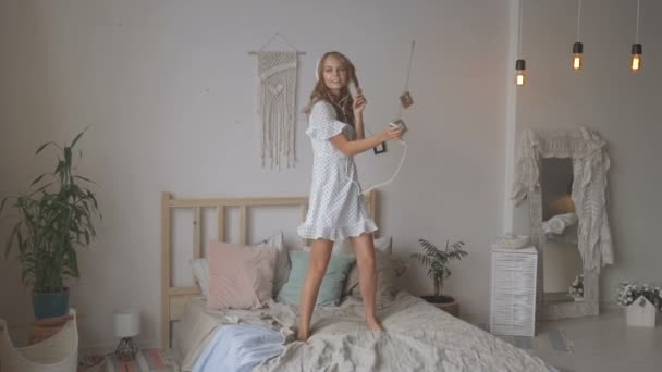 Счастливая молодая женщина в наушниках слушает музыку со смартфона и танцует на кровати дома — стоковое видео