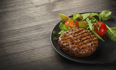 Kızarmış taze sığır burger, salata ve ahşap masada siyah tabakta otlar.