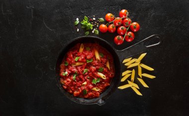 Demir döküm tavada domates sosu ve fesleğen ile makarna penne pişirme. Ahşap masa, üst görünüm.