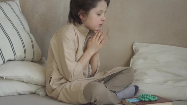 Muslim kid in prayer hat and arabic clothes with rosary beads reading holy Quran book praying to Allah, prophet Muhammad holy spirit religion symbol concept inside eastern interior — Stock Video