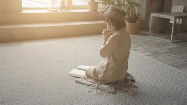 Αξιολάτρευτο μουσουλμανικό παιδί σε καπέλο προσευχής και αραβικά ρούχα με κομπολόι χάντρες ανάγνωση ιερό βιβλίο Κοράνι προσεύχεται στον Αλλάχ, προφήτης Μωάμεθ έννοια σύμβολο ιερό πνεύμα θρησκεία μέσα στο ανατολικό εσωτερικό — Αρχείο Βίντεο