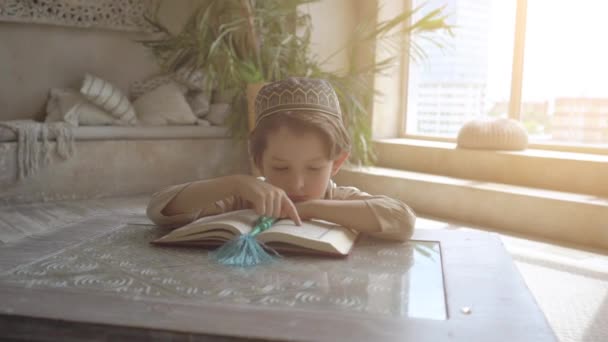 祈りの帽子とバラ色のビーズでアラビア語の服を着た6歳の少年は、東のインテリア内の預言者ムハンマドの神聖な精神宗教シンボルの概念を神に祈る聖なるクルアーンの本を読んで — ストック動画