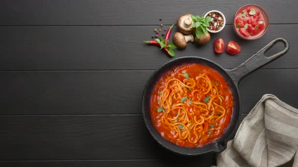 Cooking homemade Italian pasta spaghetti with tomato sauce in cast iron pan served with red chili pepper, fresh basil, cherry-tomatoes and spices over black rustic wooden background. — Stock Video