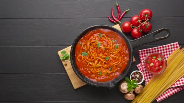 Homemade spaghetti with tomato sauce in cast iron pan served with red chili pepper, fresh basil, cherry-tomatoes and spices over black rustic wooden background, food cooking concept — Stock Video