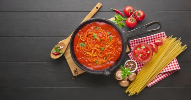 Espaguetis de pasta italiana caseros con salsa de tomate en sartén de hierro fundido servido con chile rojo, albahaca fresca, tomates cherry y especias sobre fondo de madera rústico negro, concepto de cocina de alimentos — Vídeos de Stock