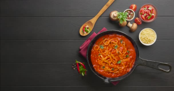 Kochen hausgemachte italienische Nudelspaghetti mit Tomatensauce in gusseiserner Pfanne, serviert mit rotem Chili, frischem Basilikum, Kirschtomaten und Gewürzen über schwarzem rustikalem Holzhintergrund. — Stockvideo