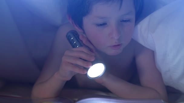 Niño curioso disfrutando leyendo interesante libro acostado debajo de una manta en la cama, sosteniendo la linterna relajándose en casa, niño feliz divirtiéndose en el dormitorio, creativo concepto de educación de la actividad infantil — Vídeo de stock