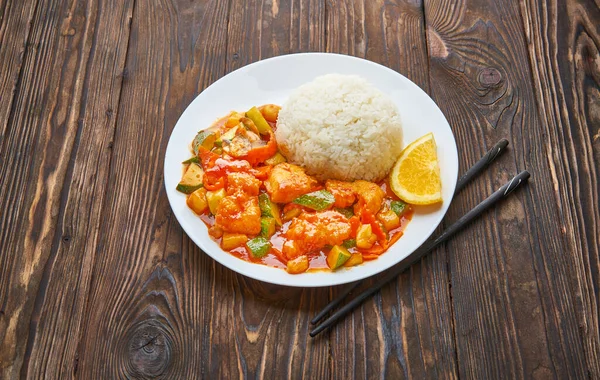 Sweet and sour chicken in orange sauce with vegetables, rice and chopsticks on wooden background, asian food top view concept with copy space