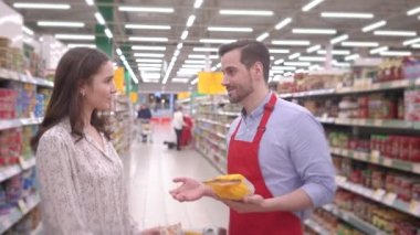 Kırmızı önlüklü arkadaş canlısı yakışıklı satıcı, genç çekici kadının süpermarkette yemek seçmesine yardımcı oluyor, müşteri ilişkileri müşteri desteği ve profesyonel işçi konsepti.
