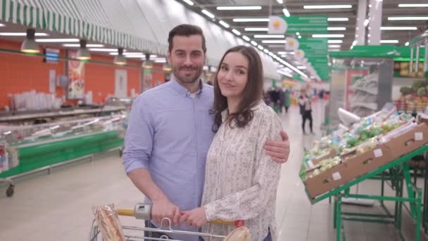 Aantrekkelijk gelukkig jong duizendjarig familie paar winkelen in supermarkt met winkelwagentje, kijken naar camera en glimlachende, mooie man en vrouw kopers klanttevredenheid supermarkt concept — Stockvideo