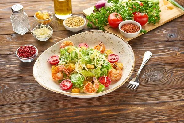 Parmesan peynirli karides sezar salatası, kızarmış ekmek, marul ve kiraz domatesleri, ahşap bir arka plan dokusunda ahşap bir tabakta çatal ve malzemeler.
