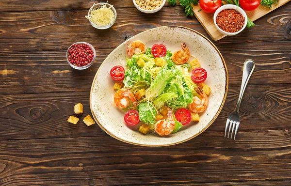 Parmesan peynirli karides sezar salatası, kızarmış ekmek, marul ve kiraz domatesleri, ahşap bir arka plan dokusunda beyaz bir tabakta çatal, malzemeli üst görünüm.