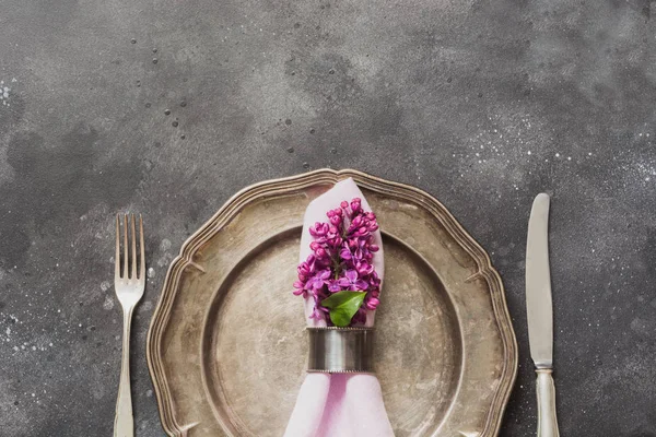 Spring elegant table place setting with violet lilac flowers, silverware on dark table. Top view.