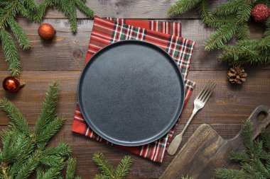 Christmas food border with black plate, evegreen branches, and red decor on wooden board. Space for text. Top view.. clipart
