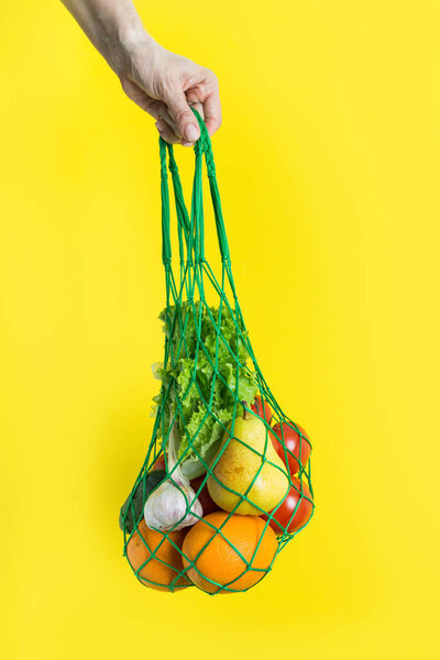 Mesh bag of fruits, vegetables, greens in woman's hand. Zero waste. Plastic free. Yellow background.