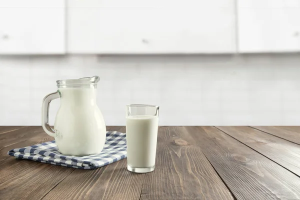 Glass of fresh milk and jug on wooden tabletop with blured white kitchen as background.