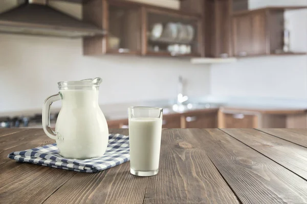 Bicchiere di latte fresco e brocca su tavolo di legno con cucina sfocata come sfondo . — Foto Stock