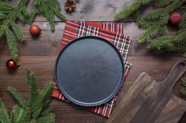 Christmas food border with black plate, evegreen branches, and red decor on wooden board. Space for text. Flat lay style. clipart