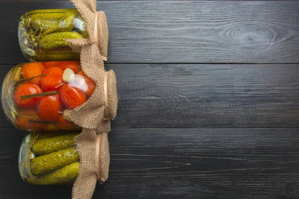 Legumbres enlatadas pepino y tomate en frascos de vidrio sobre tabla oscura de madera. Espacio para el texto. Vista desde arriba. Cosecha casera preparaciones de otoño. Tareas y tradiciones . —  Fotos de Stock