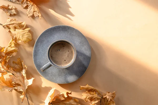 Autumn coffee in grey cup in sunny shadow and dry oak leaves on beige background. Fall mock up with copy space. View from above. Indian Summer