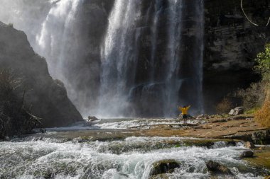 Türkiye 'deki Tortum şelalesinde dinlenen sarı ceketli bir turist. Erzurum 'daki Tortum Şelalesi' nde elleri açık bir kayanın üzerinde duran genç bir gezgin.