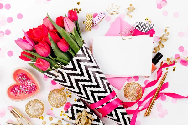 Pink and Gold Styled Desk with Florals. Pink tulips in black and white stylish wrapping paper, gifts, cosmetics and female accessories with confetti on white background