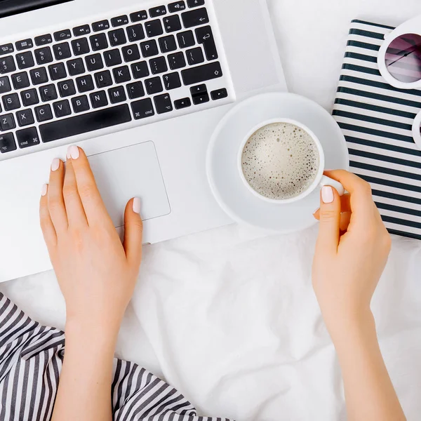 Genç bir kadın evinde, yatağında kahve içiyor ve laptopuyla çalışıyor.