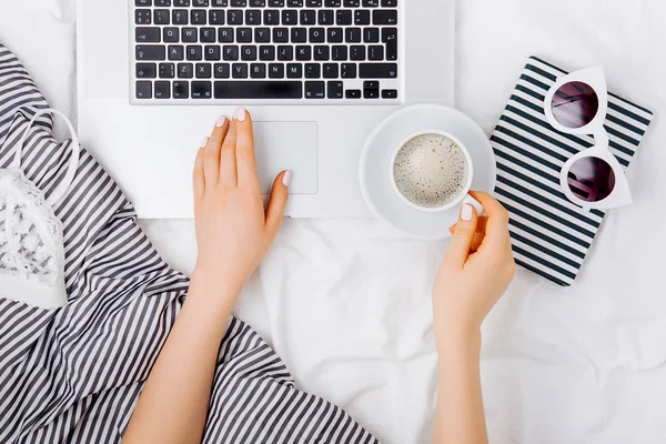 Genç bir kadın evinde, yatağında kahve içiyor ve laptopuyla çalışıyor.