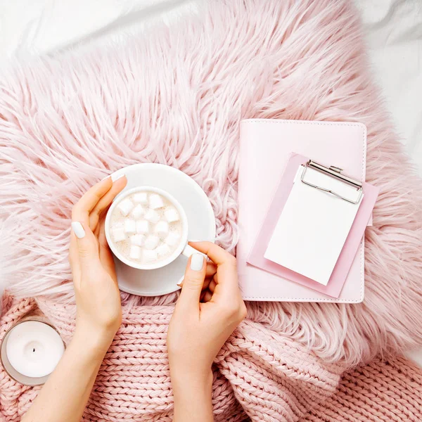 Female hands holding cup of cacao on fluffy fur plaid with clipboard and candle