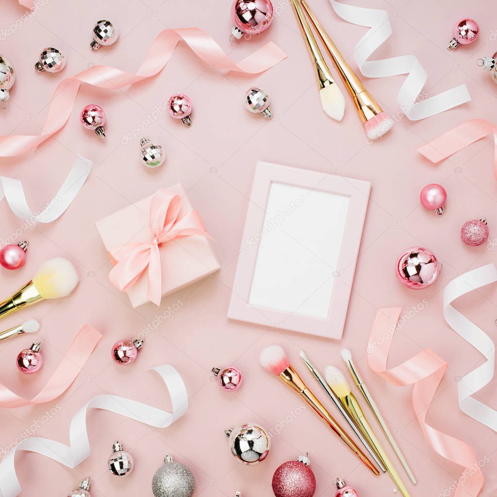 christmas baubles, ribbons, makeup brushes, gift and empty frame on pastel pink background
