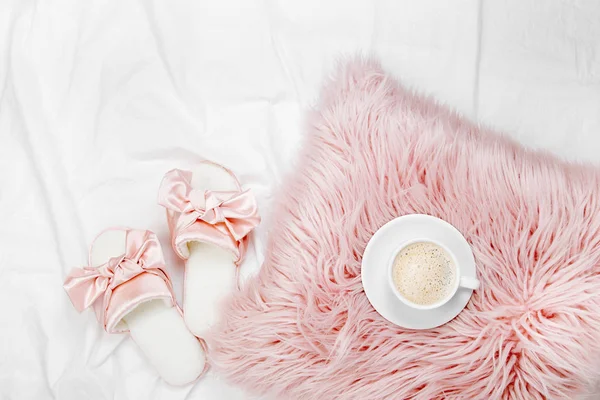 cup of coffee on pink furry pillow and slippers on bed