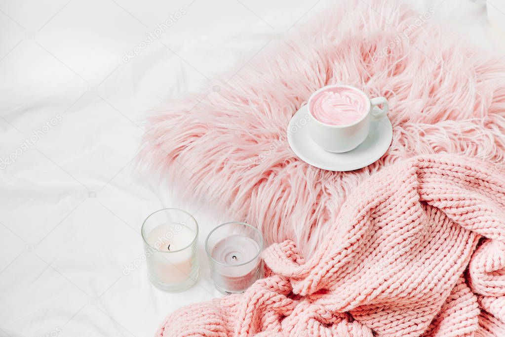 A cup of coffee on a pink pillow with candles and a blanket on the bed. 