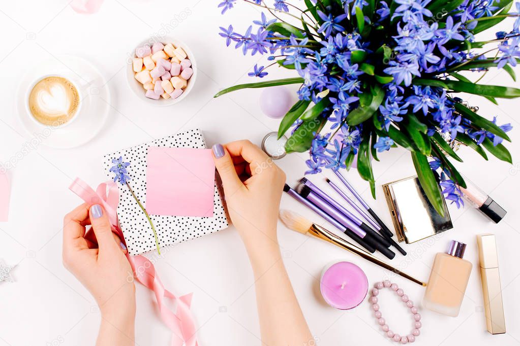 A woman unpacks a gift on background with Blue spring bouquet, coffee cup, candies and cosmetic accessories. Styled flat lay