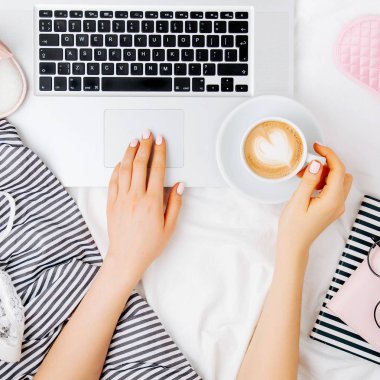 Genç bir kadın evinde, yatağında kahve içiyor ve laptopuyla çalışıyor.