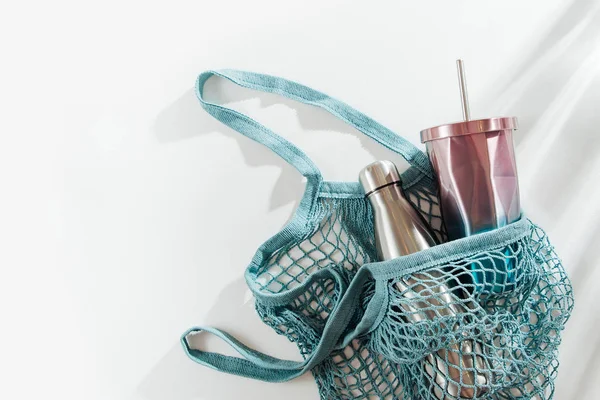 Mesh bag with reusable metal water bottle and cup on white background. Sustainable lifestyle. Zero waste concept. No plastic.