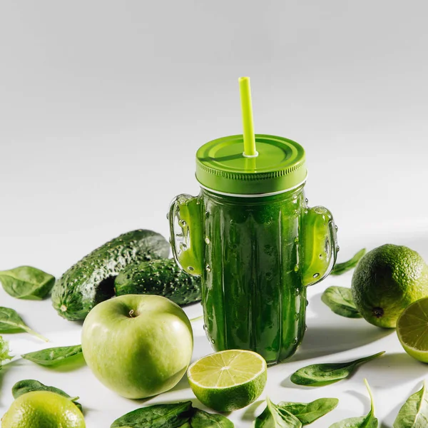 Green smoothie in cute glass jar shape of cactus with spinach and green fruits and vegetables on white table. Vegetarian food. Detox and diet concept.