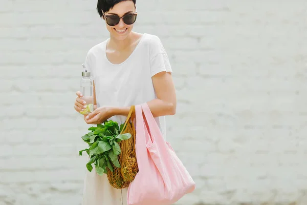Young woman in light summer clothes with a eco bag of vegetables, greens and reusable water bottle.  Sustainable lifestyle. Eco friendly concept.