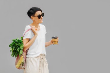 Hafif yaz kıyafetleri içinde, sebze, yeşillik ve yeniden kullanılabilir kahve kupası olan genç bir kadın. Sürdürülebilir yaşam tarzı. Çevre dostu kavram.